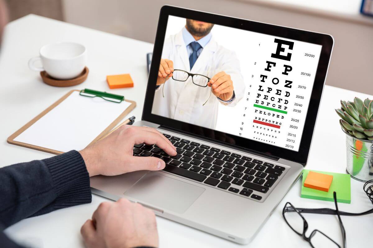 Image of an eye doctor holding a pair of glasses with an eye test next to him on a laptop screen